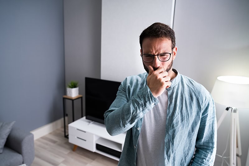 A medium shot of a bearded man with glasses standing in a living room, holding his hand up to his face and pinching his nose with a disgusted, grimacing expression as if reacting to a foul odor. He is wearing a light blue button-down shirt over a gray t-shirt, with a television console and a floor lamp visible in the background.